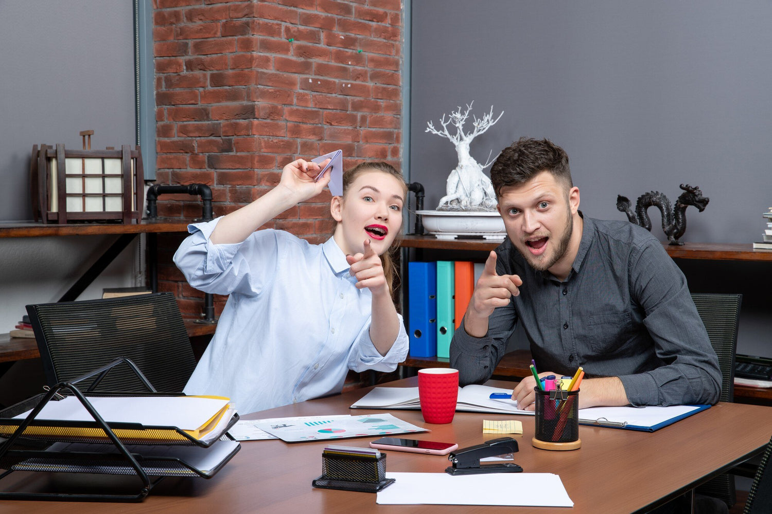 Two excited coworkers pointing at the camera in a modern office with binders and decor in the background